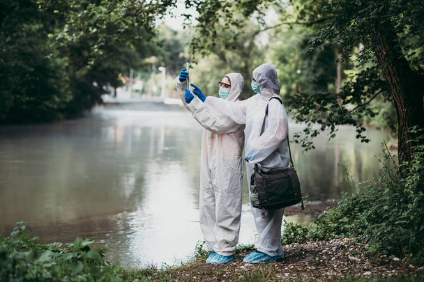 Two scientists in protective suits taking water samples from the river. - Foto, Imagen