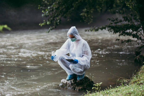 Επιστήμονας σε προστατευτικές σουίτες λαμβάνοντας δείγματα νερού από τον ποταμό. - Φωτογραφία, εικόνα