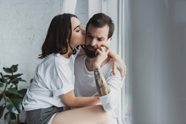 beautiful woman kissing man talking on smartphone at home - Foto, afbeelding