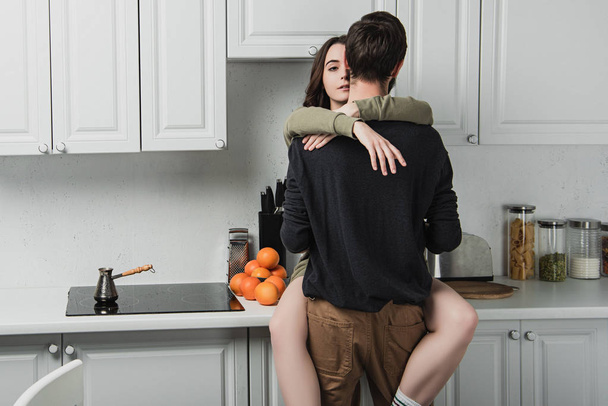 Rückansicht eines jungen Paares, das sich während des Frühstücks in der Küche umarmt - Foto, Bild