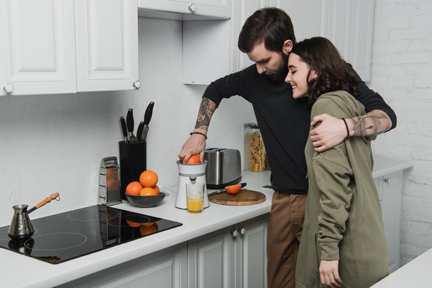 beautiful young couple making orange juice and hugging during breakfast in kitchen - Foto, imagen