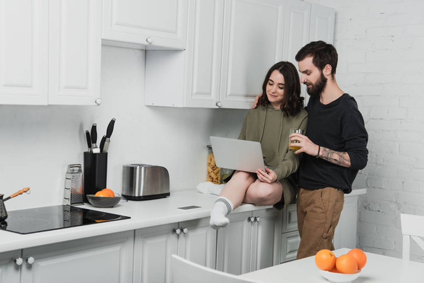 hombre guapo beber jugo mientras que la mujer usando el ordenador portátil durante el desayuno en la cocina
 - Foto, imagen