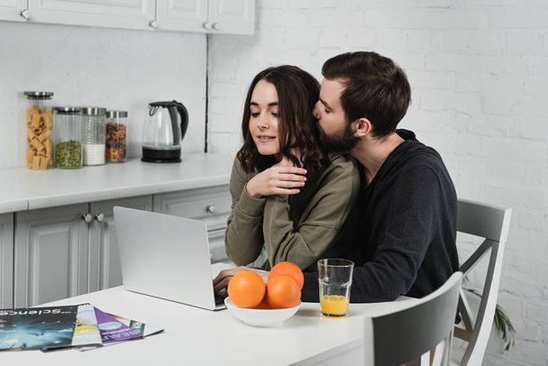 knappe man knuffelen vrouw zittend aan tafel met sinaasappels en met behulp van de laptop in de keuken - Foto, afbeelding