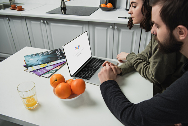 couple sitting at table and using laptop with google website on screen in kitchen - Fotografie, Obrázek