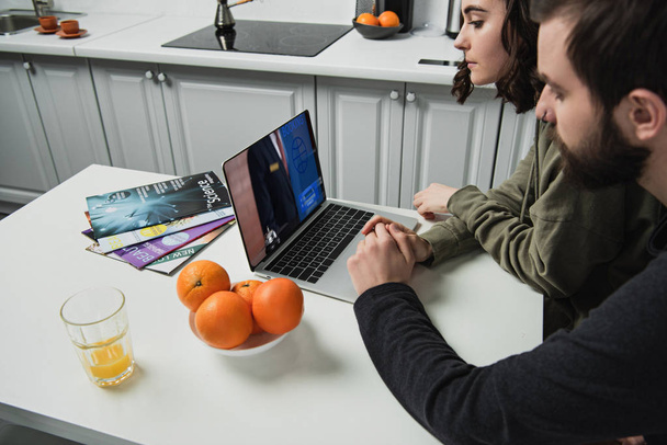 Paar sitzt am Tisch und benutzt Laptop mit Buchungswebseite auf dem Bildschirm in der Küche - Foto, Bild
