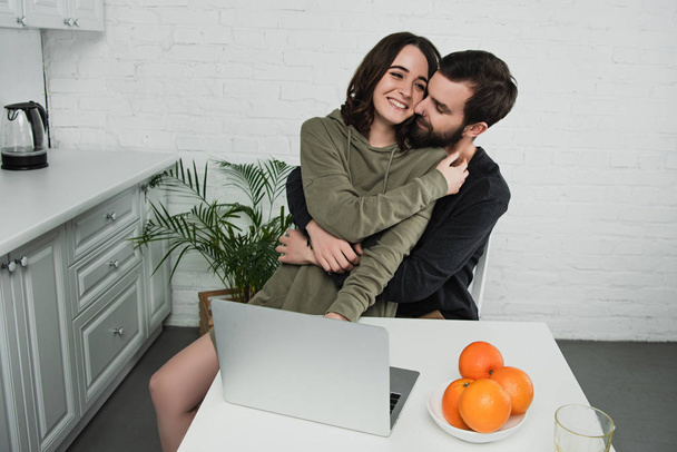 beautiful happy couple hugging while sitting at table with laptop and oranges during breakfast in kitchen - Foto, imagen