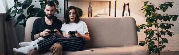beautiful surprised young couple sitting on couch with joysticks and playing video game in living room - Fotó, kép