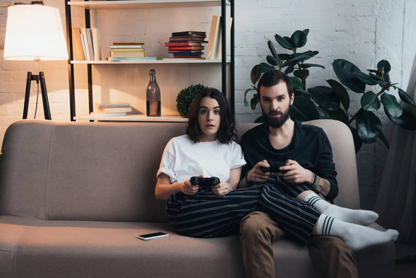 beautiful surprised young couple sitting on couch with joysticks and playing video game in living room - Foto, imagen