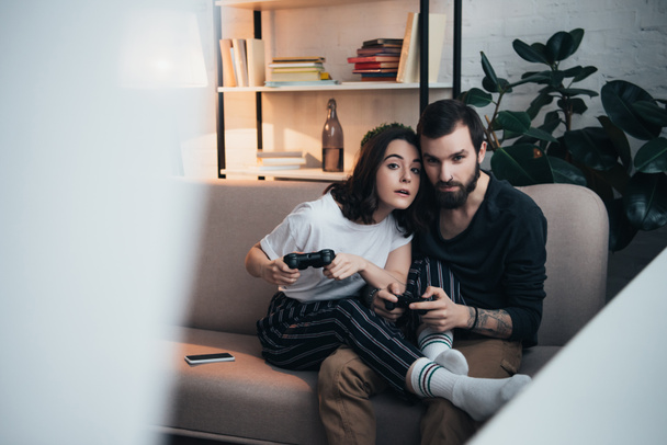 beautiful young couple sitting on couch with joysticks and playing video game in living room with copy space - Фото, изображение
