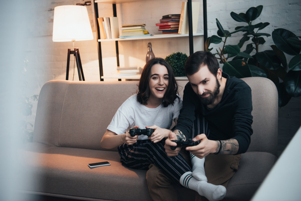 selectieve aandacht van mooie jonge paar zittend op de Bank met joysticks en afspelen van video game in woonkamer - Foto, afbeelding
