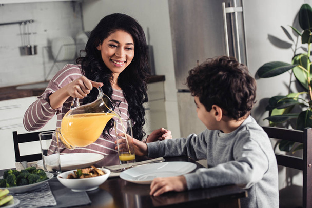 cheerful latin mother pouring orange juice into glass of son at home - Foto, immagini
