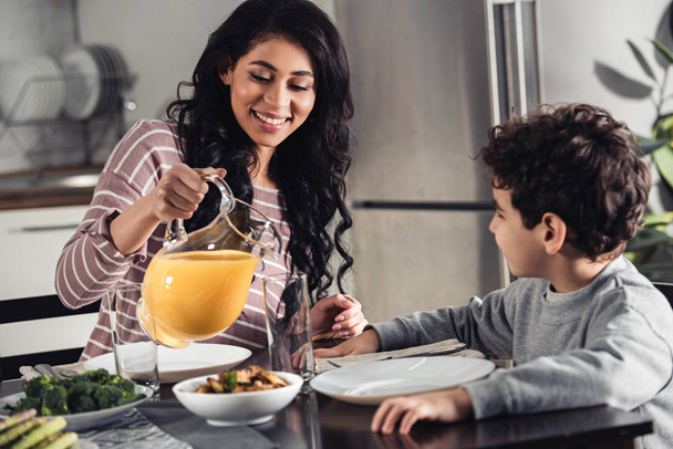 happy latin mother pouring orange juice into glass of son at home - Zdjęcie, obraz