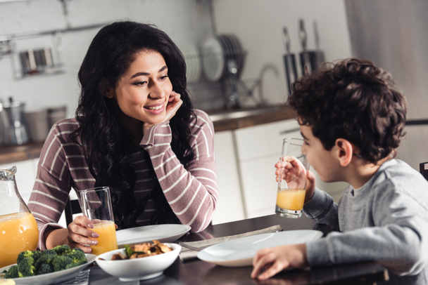 glückliche lateinische Mutter schaut Sohn an, der ein Glas Orangensaft in der Küche hält - Foto, Bild