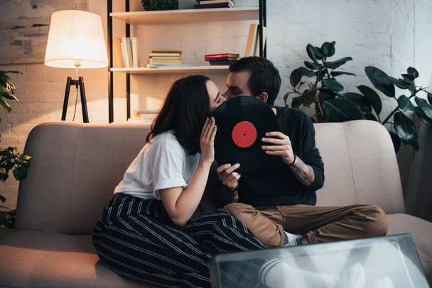 beautiful young couple covering faces with vinyl record and kissing while sitting on couch in living room - Photo, Image