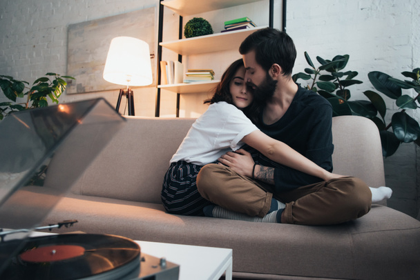 beautiful young couple sitting on couch, hugging and listening to vinyl record in living room - Фото, изображение
