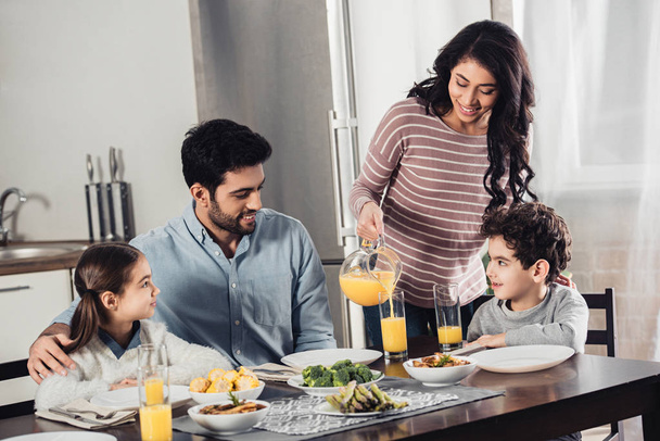 joyeuse mère latine versant du jus d'orange dans un verre de mari heureux près de la fille et du fils pendant le déjeuner
 - Photo, image