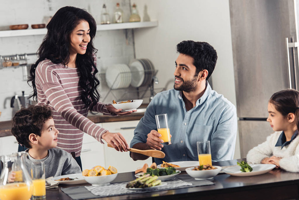 iloinen latino nainen katselee miestä pitäen lautasen lähellä tytärtä ja poikaa lounaan aikana
 - Valokuva, kuva