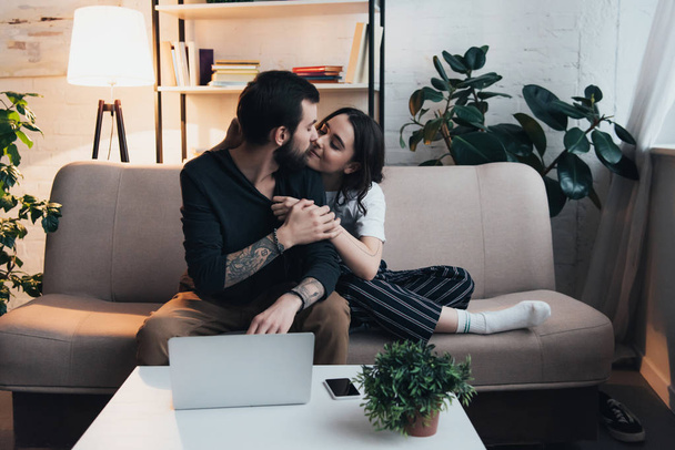 beautiful young couple sitting on couch and kissing near laptop and smartphone on table in living room - Foto, Imagem
