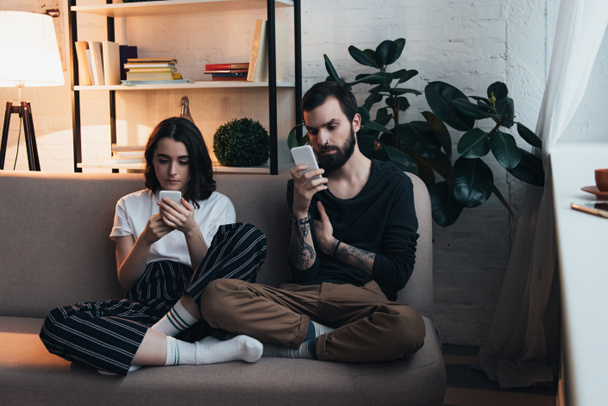 pareja joven enfocada sentado en el sofá y el uso de teléfonos inteligentes en la sala de estar
 - Foto, Imagen