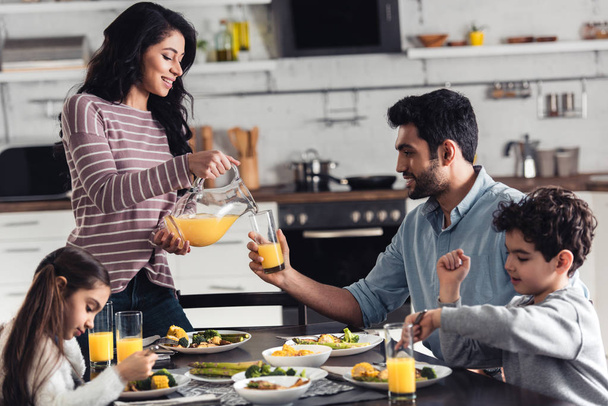 happy latin mother smiling while pouring orange juice during lunch near hispanic family - Foto, imagen