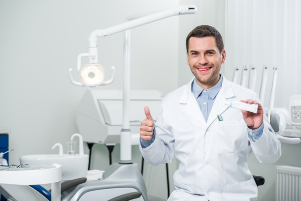 dentist holding toothpaste and showing thumb up in dental clinic - Foto, imagen