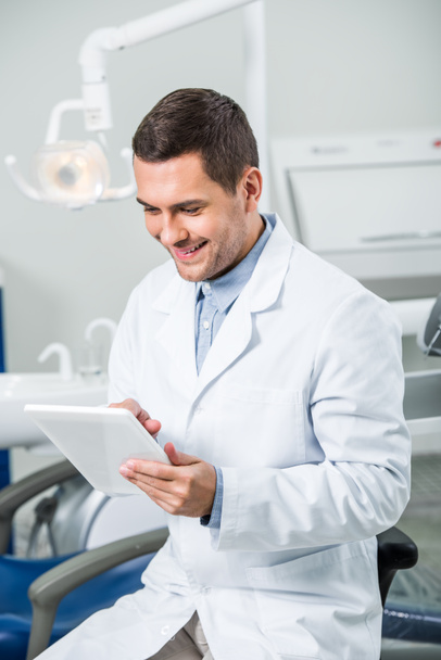 χαρούμενα οδοντίατρο σε λευκό παλτό κρατώντας ψηφιακή δισκίο σε οδοντιατρείο - Φωτογραφία, εικόνα