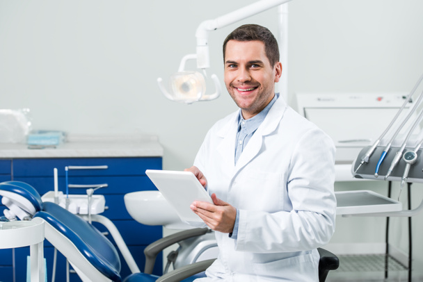 ευτυχισμένος οδοντίατρο σε λευκό παλτό κρατώντας ψηφιακή δισκίο σε οδοντιατρείο - Φωτογραφία, εικόνα