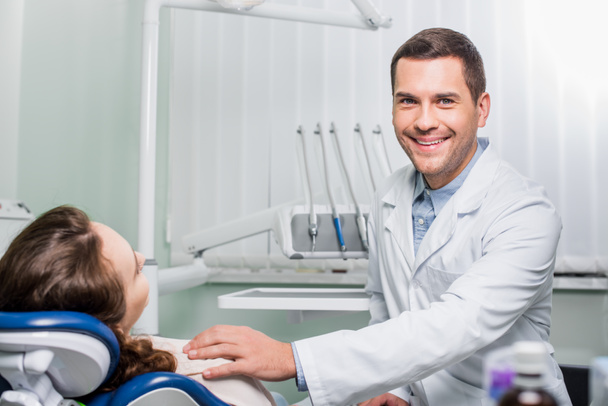 guapo dentista en blanco abrigo sonriendo cerca de mujer paciente
  - Foto, Imagen