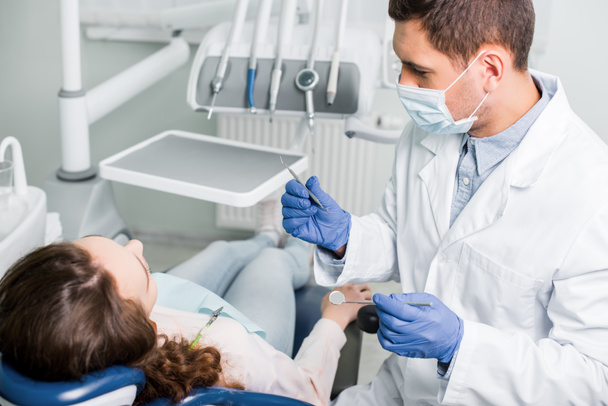 serious dentist in latex gloves and mask holding dental equipment near female patient  - Φωτογραφία, εικόνα