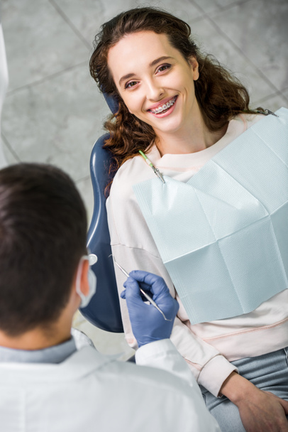 Selektivní fokus šťastná žena v závorkách během vyšetření zubů u zubaře - Fotografie, Obrázek