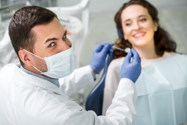 Selektivní fokus zubař v masce držící zubní zařízení během vyšetření pacientky - Fotografie, Obrázek