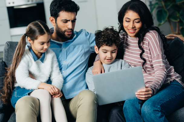 cheerful hispanic family sitting on sofa and looking at laptop at home - Foto, Bild