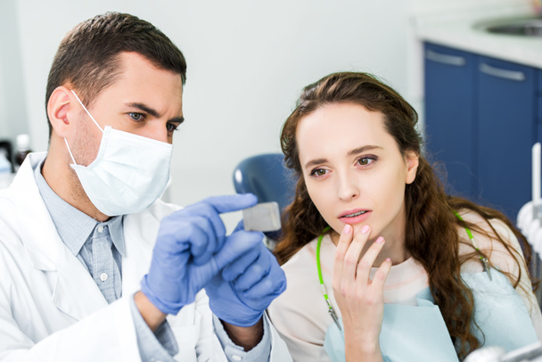 selective focus of pensive woman looking at x-ray of teeth in hands of dentist in latex gloves and mask - Photo, Image