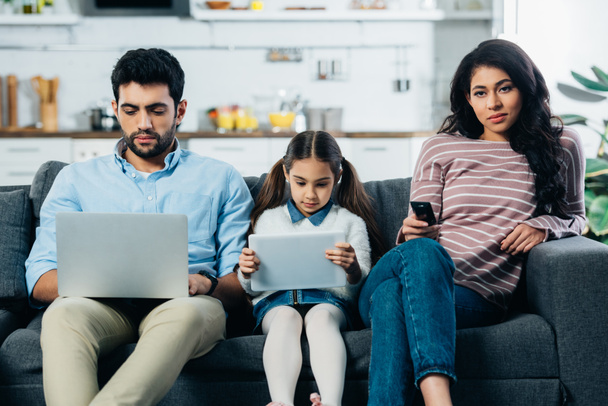 mulher latina segurando controle remoto enquanto sentado no sofá perto do marido com laptop e filha com tablet digital
  - Foto, Imagem