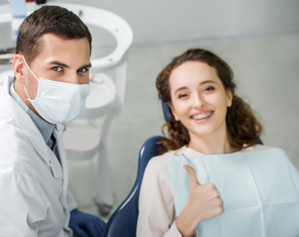 szelektív összpontosít fogorvos maszk közelében mosolyogva beteg találat hüvelykujj fel - Fotó, kép