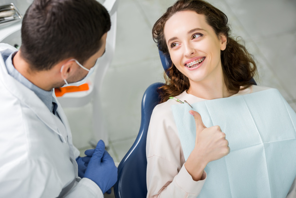 enfoque selectivo de la mujer en frenos sonriendo mientras muestra el pulgar hacia arriba cerca del dentista durante el examen
  - Foto, Imagen
