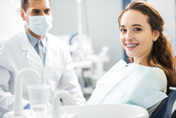 enfoque selectivo de la mujer en frenillos sonriendo con el dentista de pie en la máscara en el fondo
 - Foto, Imagen