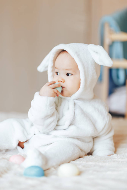 kleines süßes Kind lächelt fröhliches Baby in einem weißen Hasen Hasenkostüm Ostern spielen mit bunten Ostereiern wollte sie essen - Foto, Bild