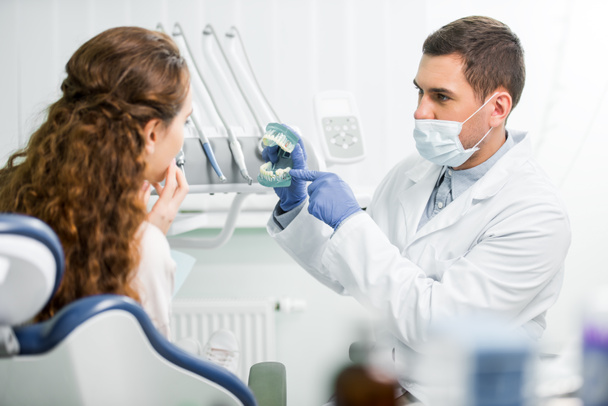 selectieve aandacht van tandarts in masker en latex handschoenen wijzen met de vinger naar model van de tanden in de buurt van vrouwelijke patiënt - Foto, afbeelding