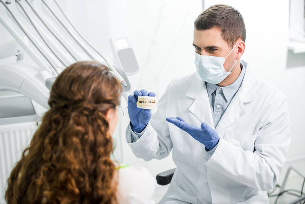 selective focus of dentist in mask and latex gloves showing teeth model near woman - Foto, Imagen