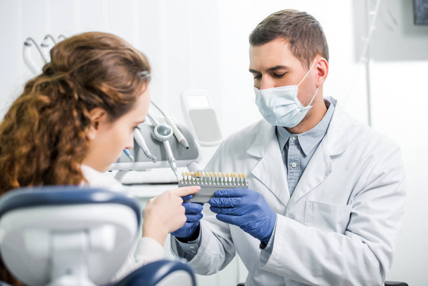 mujer señalando con el dedo a los dientes paleta de colores cerca del dentista en la máscara y guantes de látex
 - Foto, Imagen