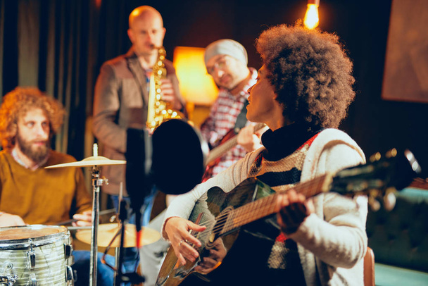 Mixed race woman singing and playing guitar while sitting on chair.In background drummer, saxophonist and bass guitarist. - Photo, Image