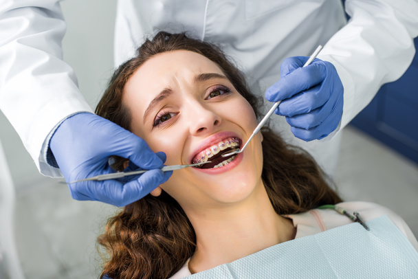 vista recortada del dentista en guantes de látex examinando a mujer atractiva en frenos con la boca abierta
  - Foto, Imagen