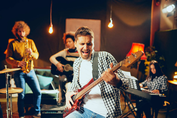 Mann singt und spielt Bassgitarre, während er im heimischen Studio sitzt. Im Hintergrund spielt seine Band Instrumente. - Foto, Bild