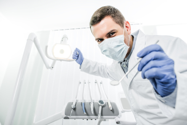 dentist in latex gloves and mask holding dental lamp and dental instrument in hands - Foto, Imagen