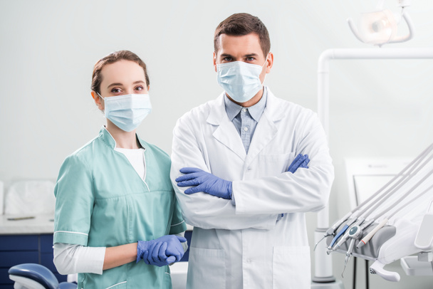dentista donna in maschera in piedi vicino a collega con le braccia incrociate in clinica dentale
 - Foto, immagini