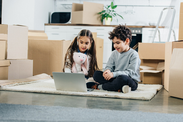 enfants heureux en utilisant un ordinateur portable tout en étant assis sur le tapis dans la nouvelle maison
 - Photo, image