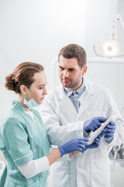 male dentist holding digital tablet and looking at attractive colleague in dental clinic - Photo, Image