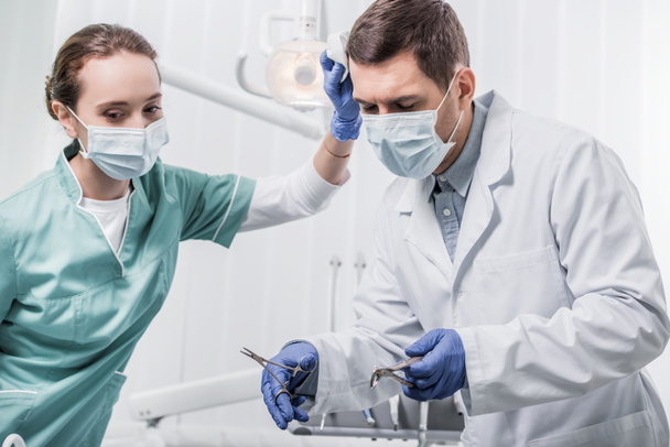 dentista mujer en máscara de pie cerca de colega que sostiene instrumentos dentales
  - Foto, Imagen