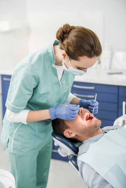 dentista femenina con máscara y guantes de látex examinando paciente con la boca abierta
 - Foto, Imagen